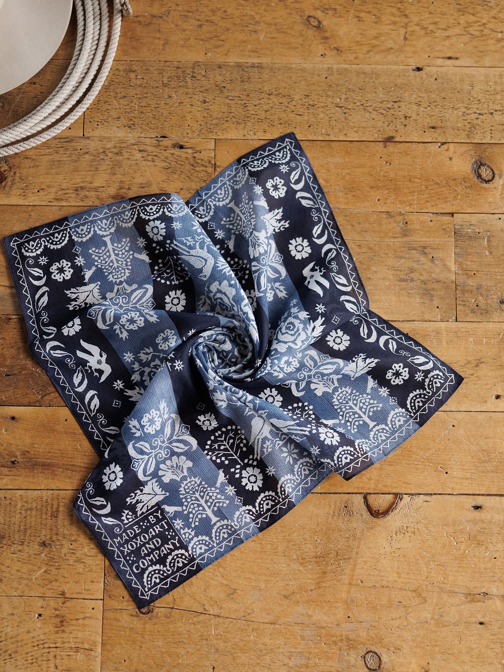 The Coverlet Western Floral Cotton-Silk Neck Scarf by Savannah Sevens western life{&}style, featuring blue and white floral and animal patterns, is displayed on rustic wood with a coiled rope partially visible in the top left corner.