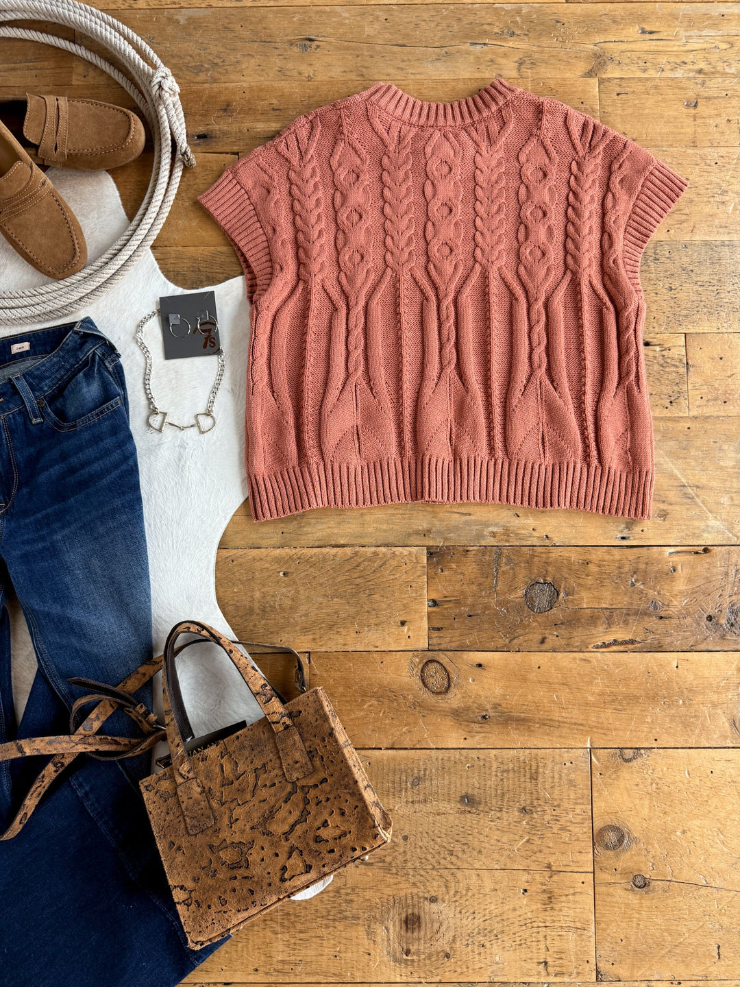 A Covina Burnt Sienna Knit Top by Savannah Sevens western life{&}style, blue jeans, tan loafers, a cork handbag, a silver bracelet, and a white blanket are arranged on a wooden floor.