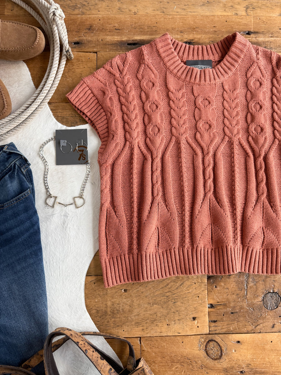 The Covina Burnt Sienna Knit Top by Savannah Sevens western life{&}style is displayed on a wooden surface alongside blue jeans, a silver necklace, a brown shoe, and a brown leather bag. Its relaxed fit perfectly complements this casual ensemble.