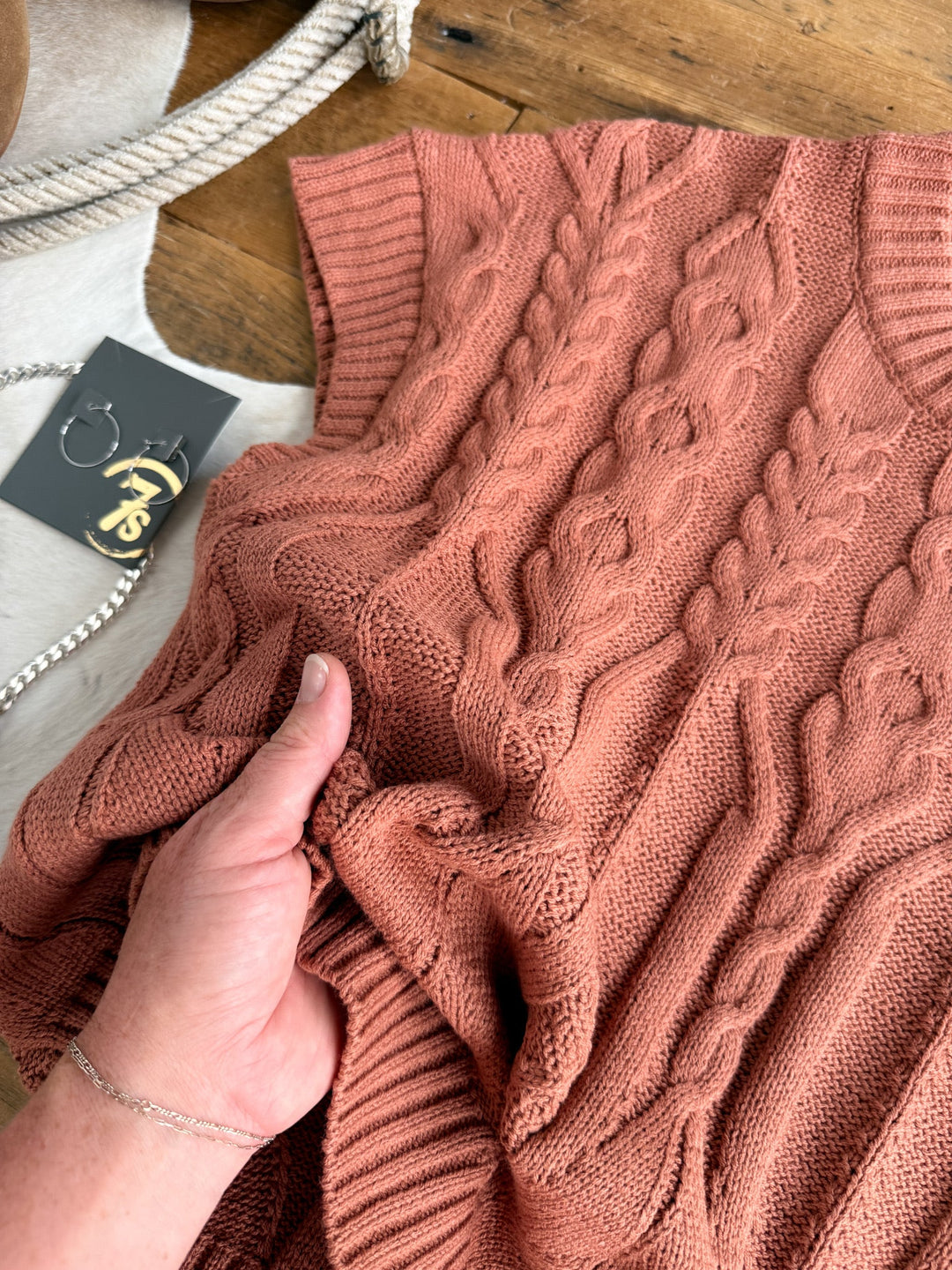 A hand holds the Covina Burnt Sienna Knit Top by Savannah Sevens western life{&}style, a pinkish-brown short sleeve knit with a relaxed fit, displayed on a wooden surface beside a jewelry tag and rope.