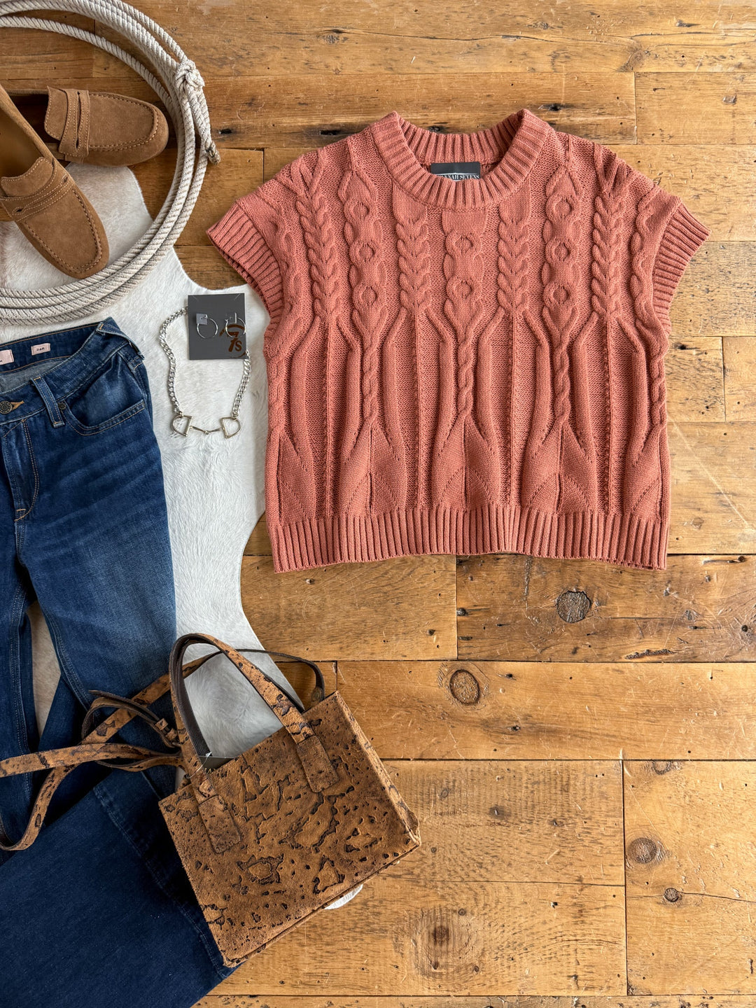 A flat lay featuring the Covina Burnt Sienna Knit Top by Savannah Sevens western life{&}style, styled with blue jeans, tan loafers, a cork handbag, a white bracelet, a coiled rope, and a white rug on a wooden floor.