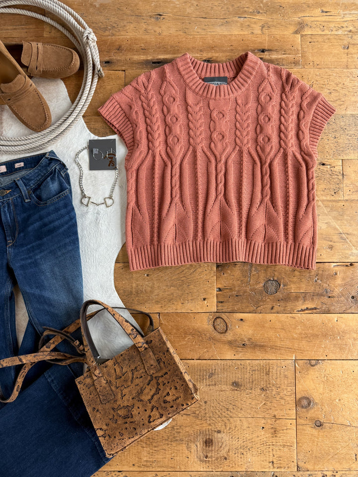 A flat lay featuring the Covina Burnt Sienna Knit Top by Savannah Sevens western life{&}style, styled with blue jeans, tan loafers, a cork handbag, a white bracelet, a coiled rope, and a white rug on a wooden floor.
