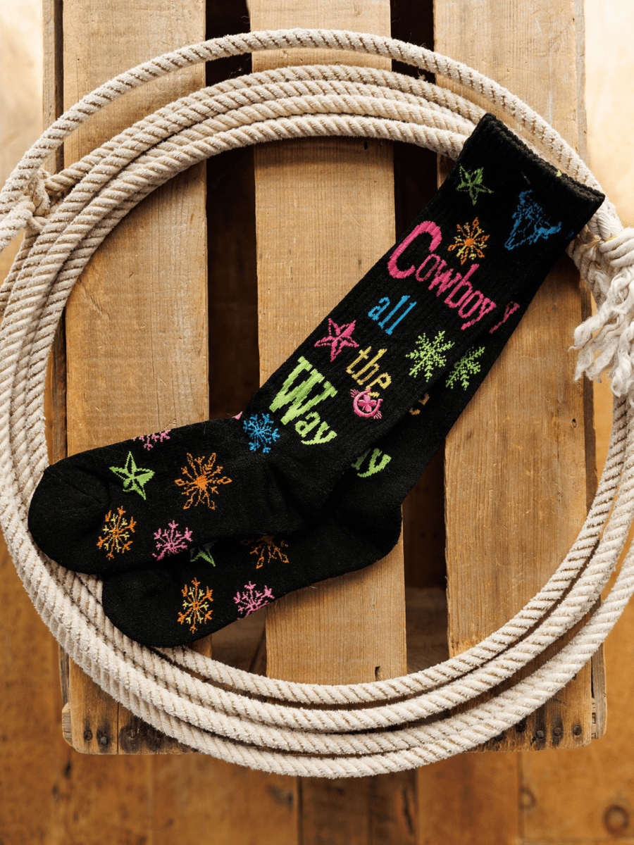 The Cowboy All The Way Crew Socks by Savannah Sevens western life{&}style feature colorful star and snowflake patterns with festive text, an odor-fighting design, and are shown on a wooden surface framed by a coiled white rope.