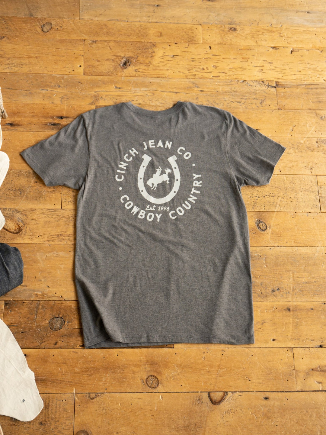 A Cowboy Country Men's Graphic Tee by Savannah Sevens western life{&}style lies flat on a wooden floor, showing a white graphic on the back with a horseshoe, cowboy riding a bucking horse, and "Cinch Jean Co. Cowboy Country Est. 1996.