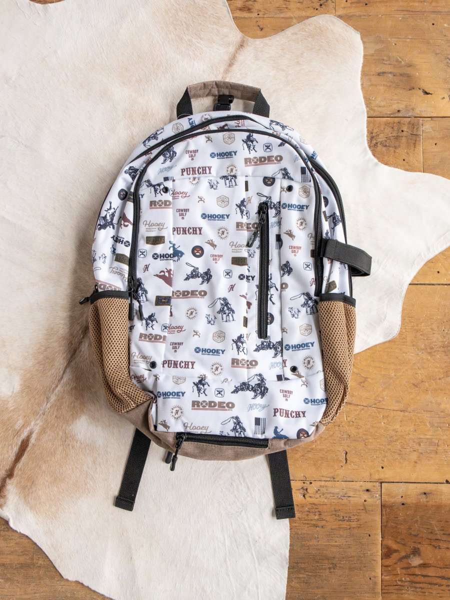 The Cowboy Golf Medium Canvas Hat Holder Backpack by Savannah Sevens western life{&}style features cowboy-themed designs, tan mesh side pockets, and a Hooey hat holder, shown on a cowhide rug atop a wooden floor.
