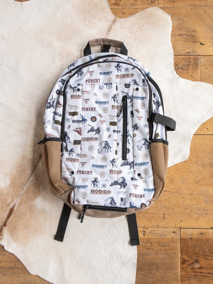 The Cowboy Golf Medium Canvas Hat Holder Backpack by Savannah Sevens western life{&}style features cowboy-themed designs, tan mesh side pockets, and a Hooey hat holder, shown on a cowhide rug atop a wooden floor.