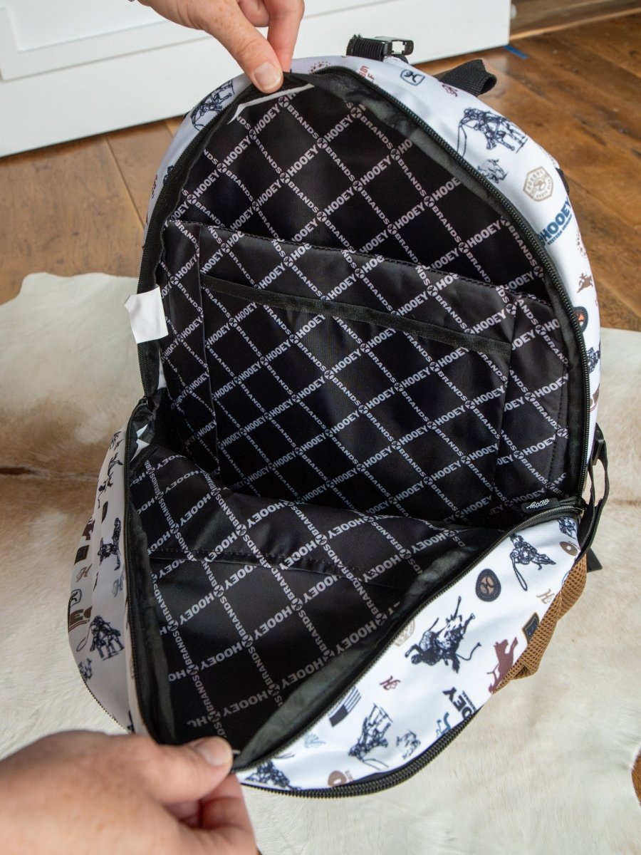 A person opens a Cowboy Golf Medium Canvas Hat Holder Backpack by Savannah Sevens western life{&}style, revealing HOOEY-patterned lining. The white exterior has black and blue illustrations and features a Hooey hat holder. Scene: indoors on wood floor and rug.