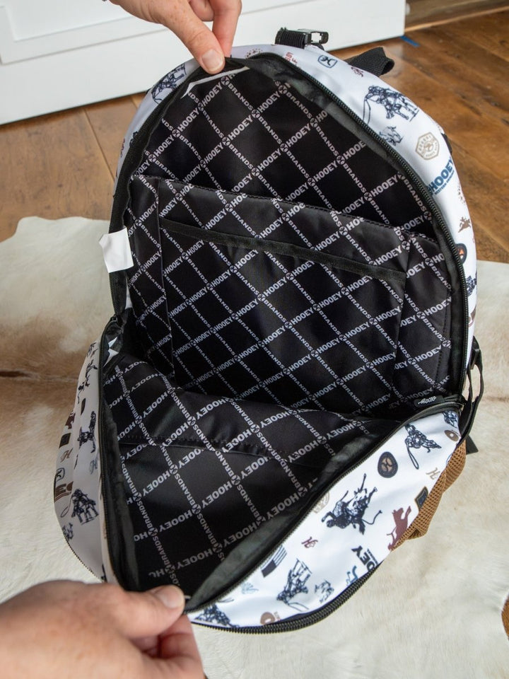 A person opens a Cowboy Golf Medium Canvas Hat Holder Backpack by Savannah Sevens western life{&}style, revealing HOOEY-patterned lining. The white exterior has black and blue illustrations and features a Hooey hat holder. Scene: indoors on wood floor and rug.