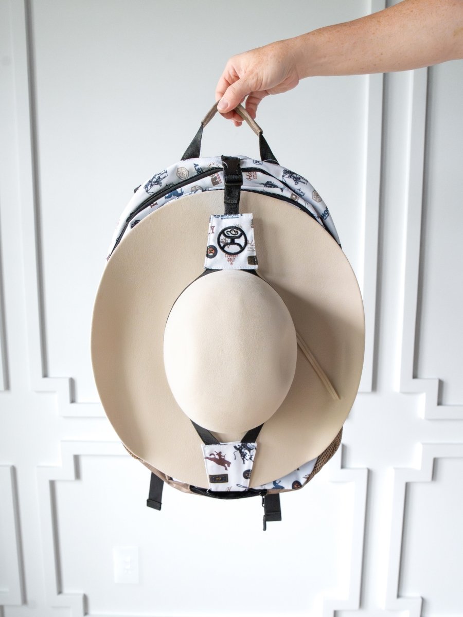 A hand holds the Savannah Sevens western life{&}style Cowboy Golf Medium Canvas Hat Holder Backpack with a large beige hat attached. A white wall with decorative molding appears in the background.