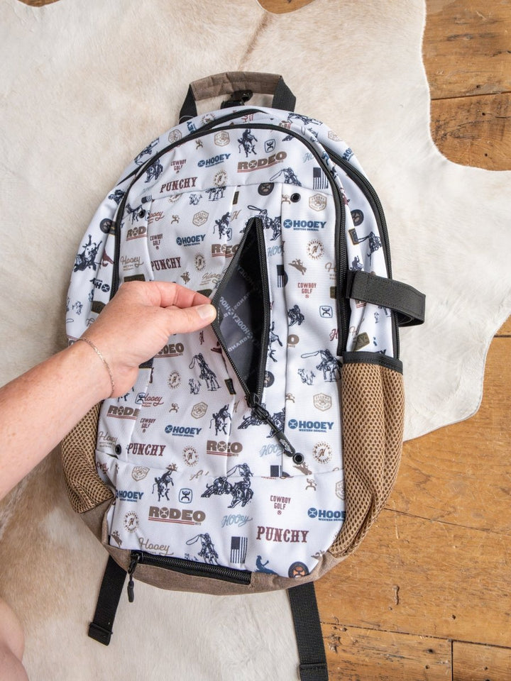 A hand opens a hidden zipper pocket on the Savannah Sevens western life{&}style Cowboy Golf Medium Canvas Hat Holder Backpack, adorned with cowboy golf graphics and brown trim, resting on a cowhide rug over a wooden floor.