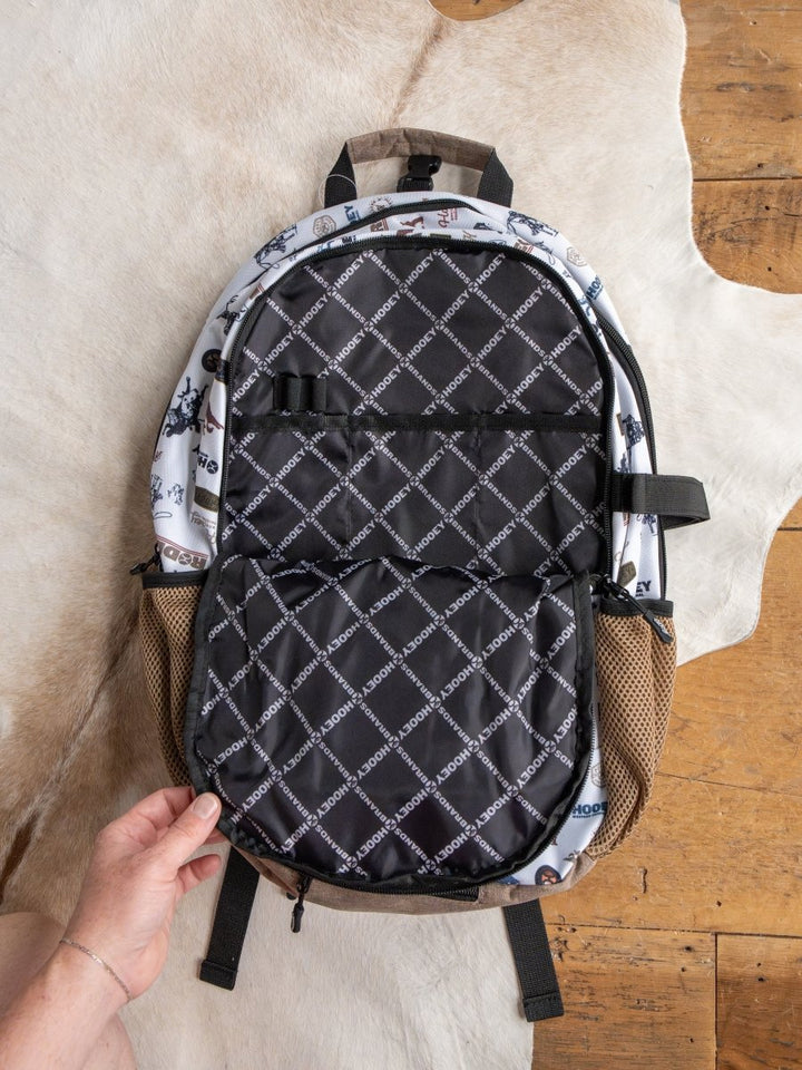 A hand holds the Cowboy Golf Medium Canvas Hat Holder Backpack by Savannah Sevens western life{&}style, shown open on a cowhide rug and wood floor. It features a black and white patterned interior, mesh side pockets, brown base, and Hooey hat holder.