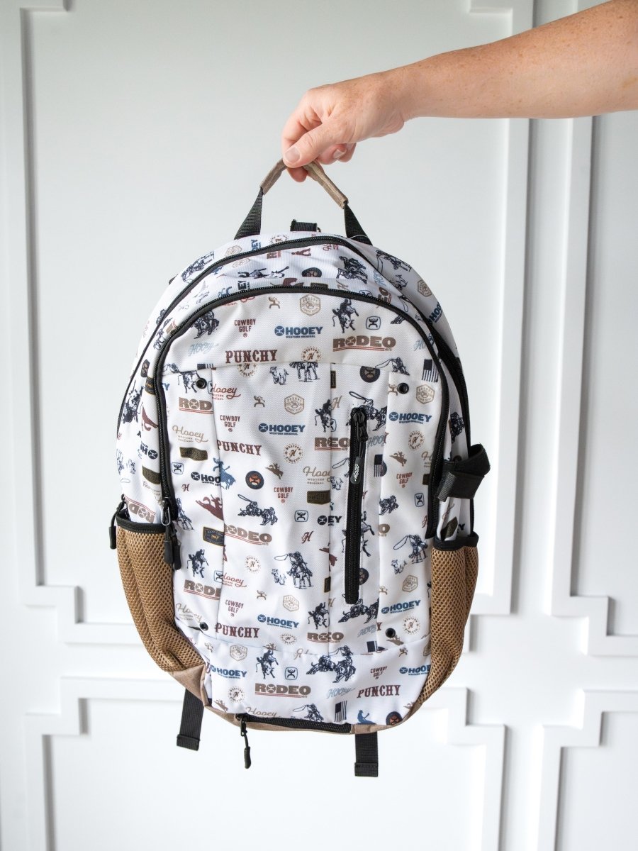 A hand displays the Cowboy Golf Medium Canvas Hat Holder Backpack by Savannah Sevens western life{&}style, featuring cowboy golf graphics and brown mesh side pockets, against a white paneled wall background.