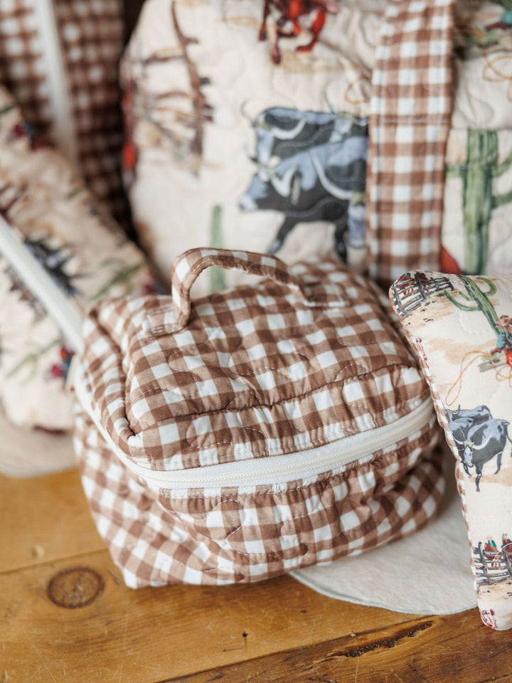 A small quilted bag with a brown and white vintage gingham pattern, white zipper, and handle from the Cowgirl Gingham Collection by Savannah Sevens western life&style sits on a wooden surface next to other quilted travel bags.
