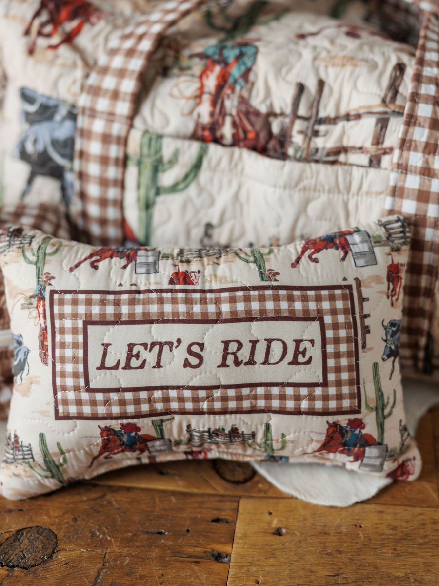 A small decorative pillow with a brown plaid border and "LETS RIDE" sits on wood before a cowboy-themed quilt—perfectly complementing the Cowgirl Gingham {Collection} by Savannah Sevens western life{&}style.