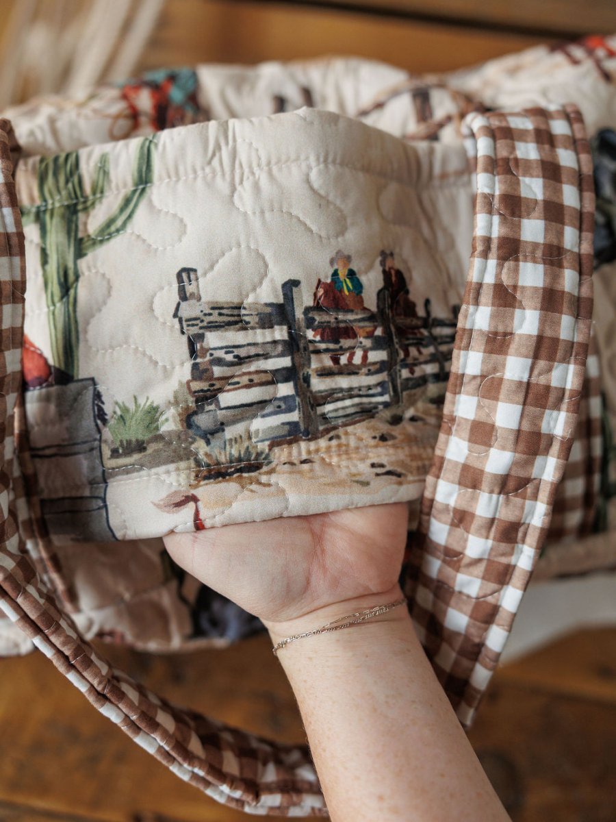 A hand holds a Cowgirl Gingham Collection travel bag by Savannah Sevens western life{&}style, featuring a vintage stitched cowboy scene with two on horseback near a fence and cactus in beige, brown, and green tones.