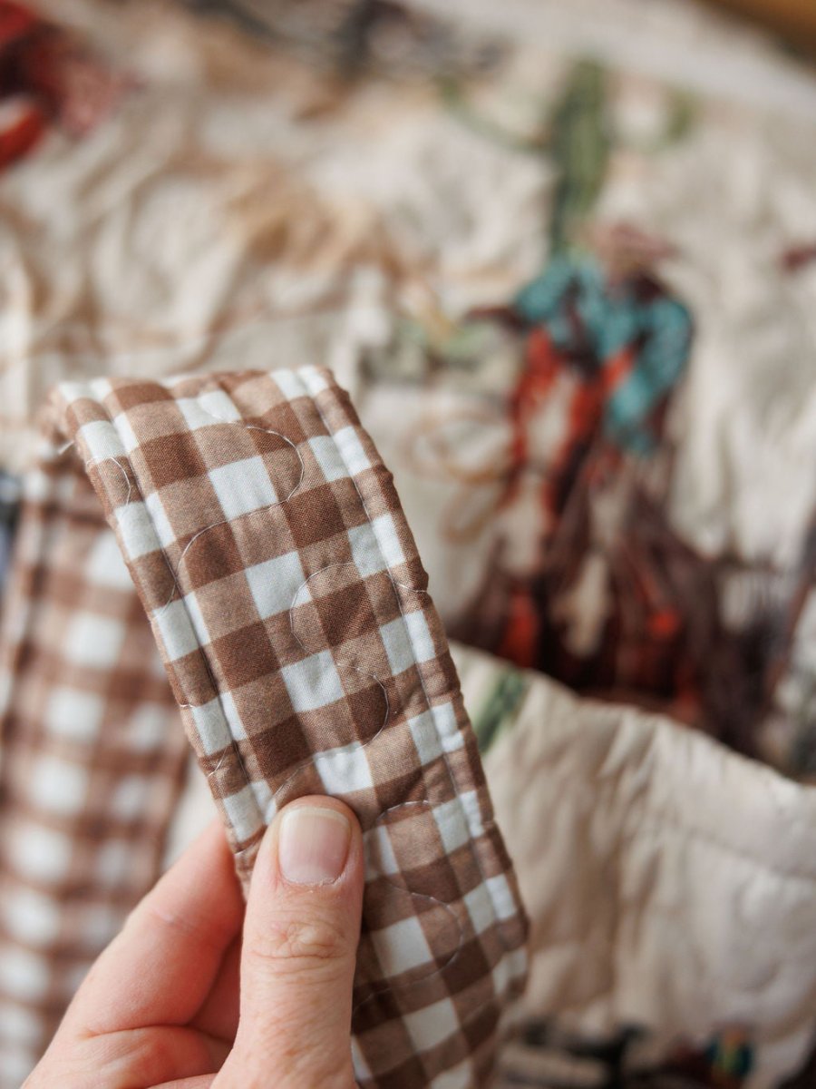A hand holds the edge of a Cowgirl Gingham {Collection} quilt by Savannah Sevens western life{&}style, with brown and white gingham and a blurred background showing a colorful cowboy design inspired by western bag collections.