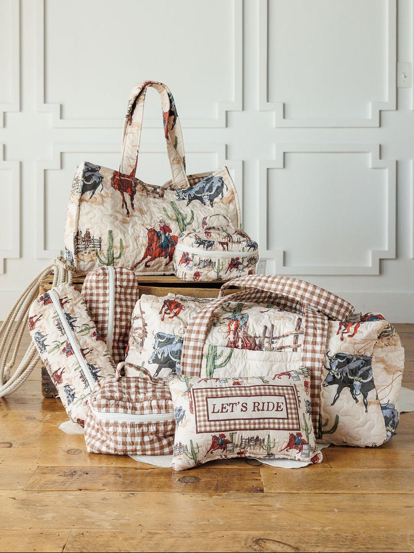 The Cowgirl Gingham Collection by Savannah Sevens western life{&}style features quilted travel bags and a “LET’S RIDE” pillow with cowboy, horse, cactus, and vintage gingham prints, arranged on a wooden floor against a white paneled wall.