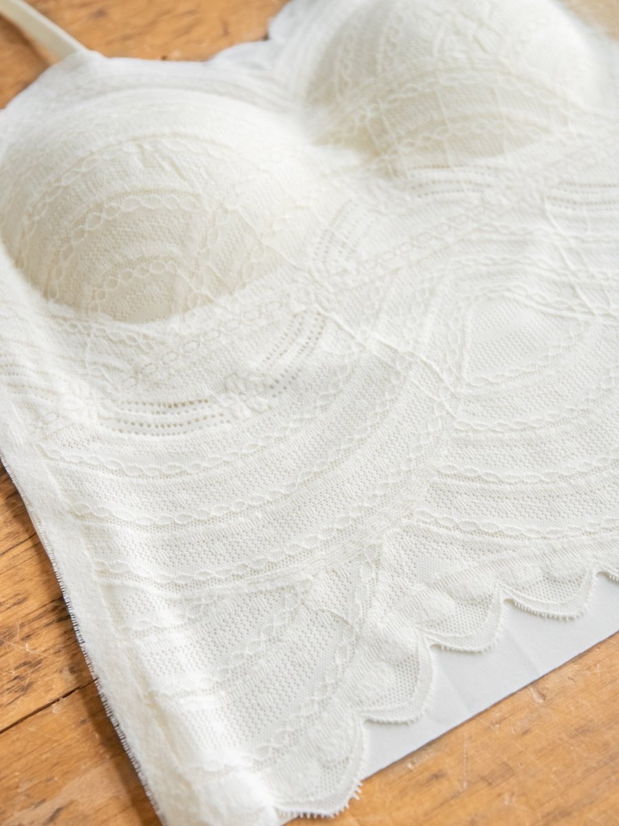 A close-up of the Creston Bralette by Savannah Sevens western life{&}style, featuring cream-colored padded cups with intricate lace and scalloped edges, displayed flat on a wooden surface.