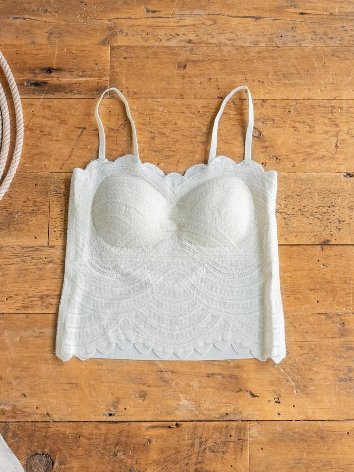 The Creston Bralette by Savannah Sevens western life{&}style, featuring white lace with scalloped edges, adjustable straps, and padded cups, is displayed flat on a wooden surface.