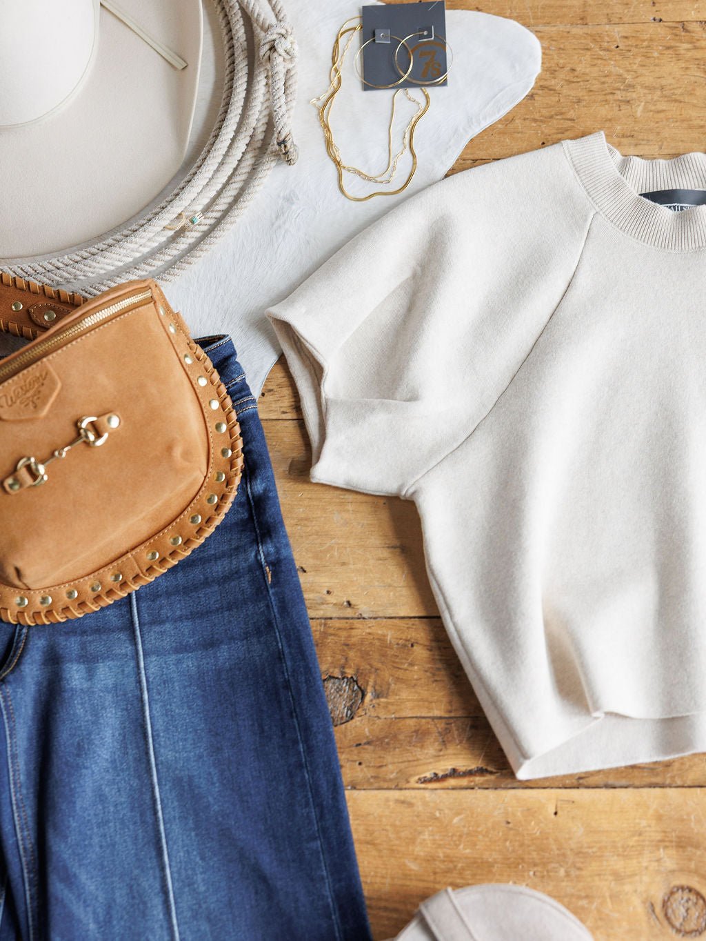 A flat lay showcases the Crestview Oatmeal Top by Savannah Sevens western life{&}style, styled with blue jeans, a tan suede crossbody bag, a white hat, gold jewelry, and a white rope on a wooden surface.