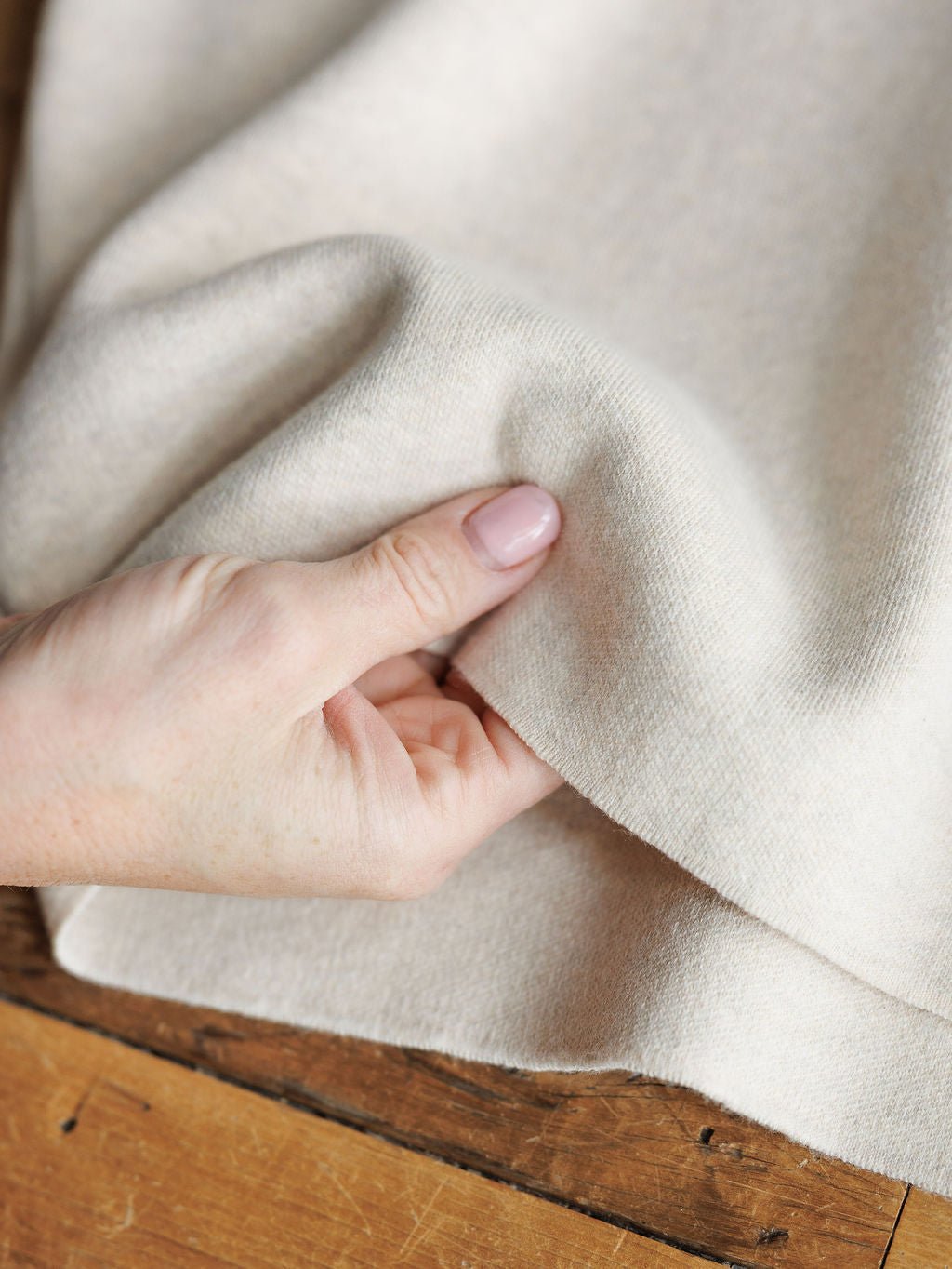 A close-up shows a hand with light pink nails gently lifting the Crestview Oatmeal Top by Savannah Sevens western life{&}style—a soft, cream-colored short sleeve top with a relaxed fit, displayed on a wooden surface.