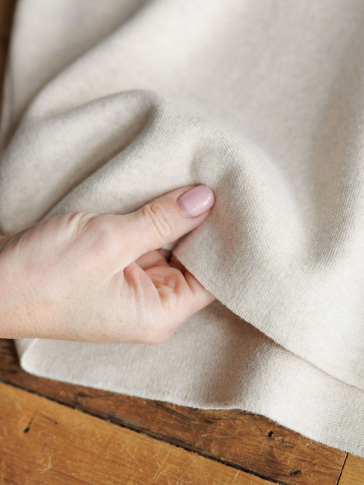 A close-up shows a hand with light pink nails gently lifting the Crestview Oatmeal Top by Savannah Sevens western life{&}style—a soft, cream-colored short sleeve top with a relaxed fit, displayed on a wooden surface.