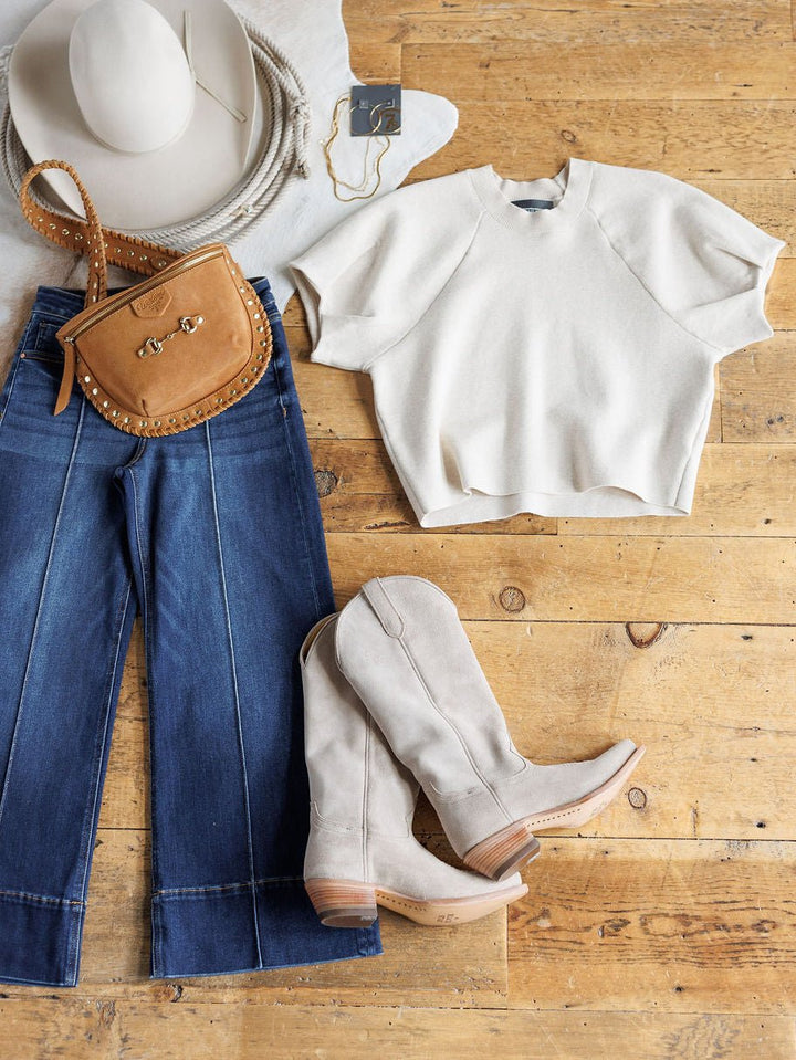 Flat lay of a relaxed outfit featuring the Crestview Oatmeal Top by Savannah Sevens western life{&}style, blue wide-leg jeans, beige cowboy boots, tan studded crossbody bag, layered necklaces, and a white hat on a wooden floor.