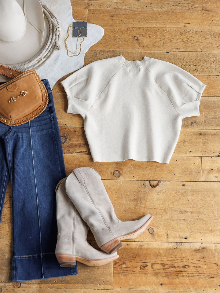 Flat lay of a casual outfit on wood, featuring blue relaxed-fit jeans, Savannah Sevens western life{&}style Crestview Oatmeal Top, tan suede cowboy boots, tan crossbody bag, white hat, and gold jewelry.