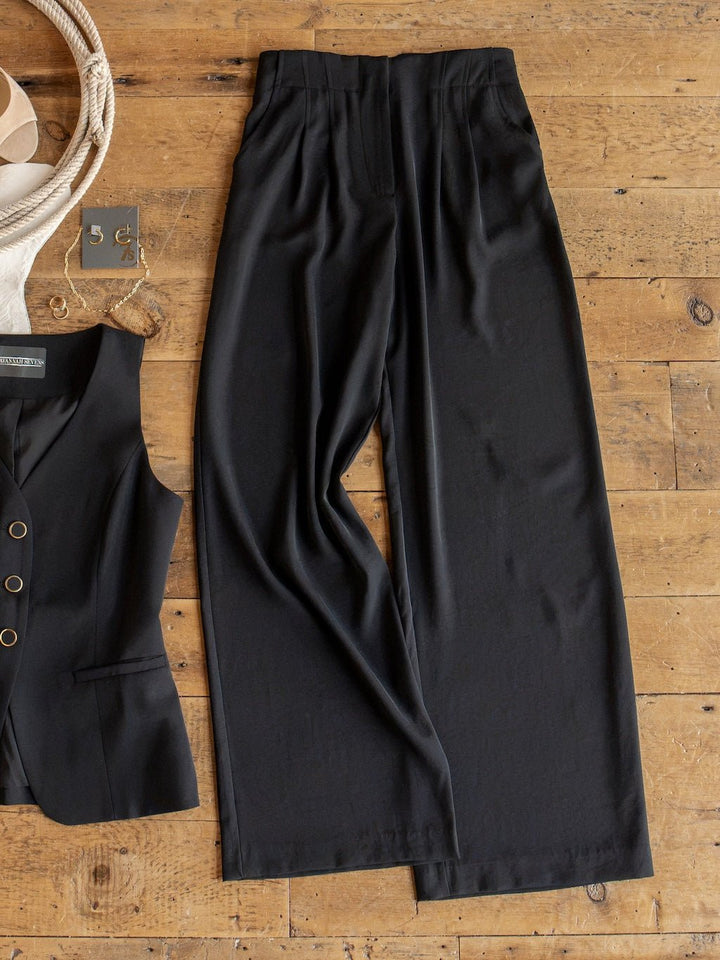 The Cuero Black Vest Set by Savannah Sevens western life{&}style is displayed laid flat on a wooden floor, featuring the matte satin wide-leg black pants alongside the matching black vest top. Accessories including a necklace and earrings are arranged in the corner, while the high-waisted wide-leg pants complete this sophisticated look.