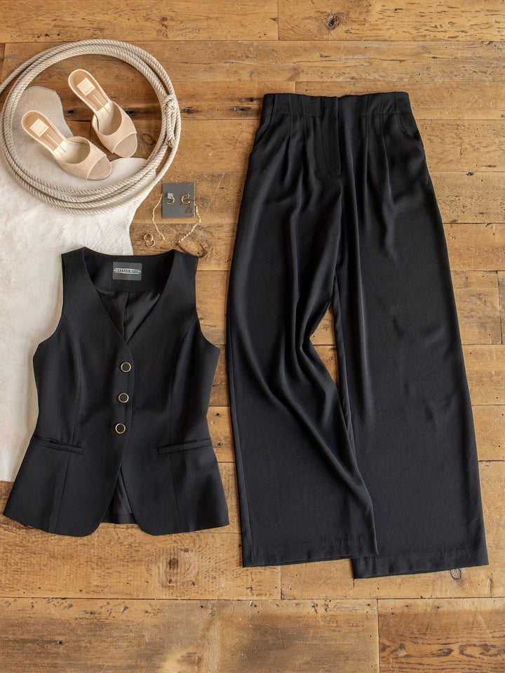 The Cuero Black Vest Set from Savannah Sevens western life{&}style—a black sleeveless vest with gold buttons and matching high waisted, wide leg pants in matte satin—is laid out on a wooden floor alongside beige sandals, a woven bag, and gold earrings.