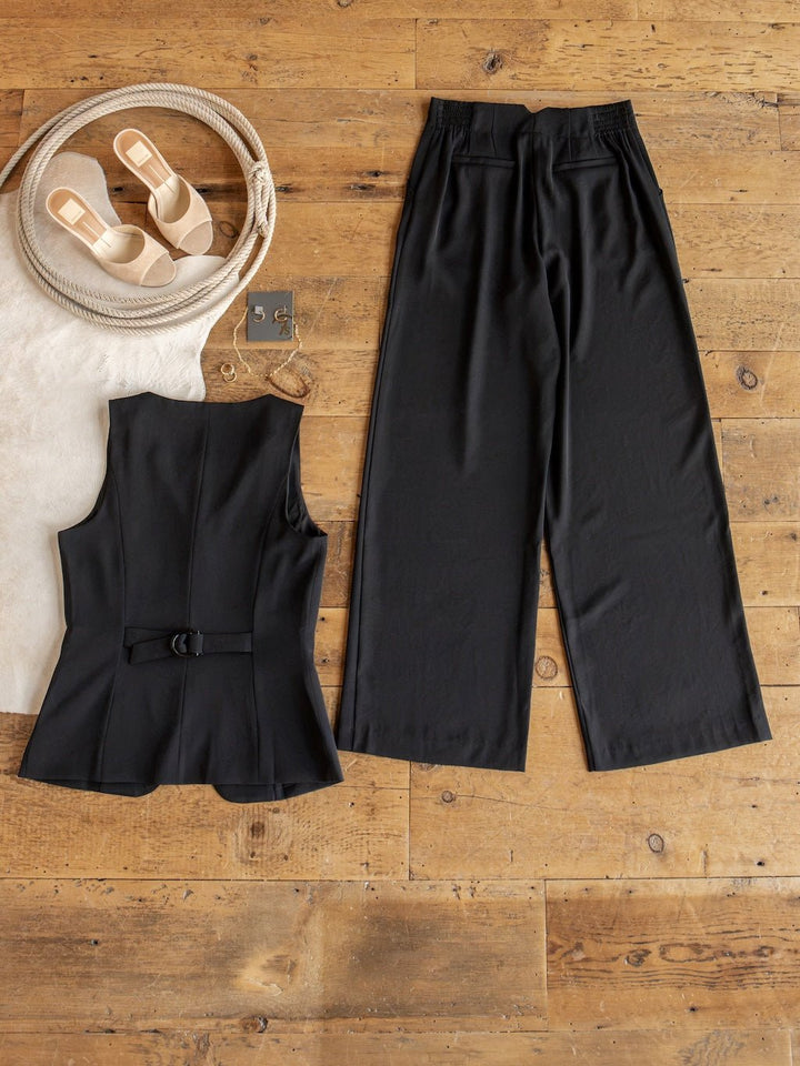 The Cuero Black Vest Set by Savannah Sevens western life{&}style, featuring a sleeveless belted matte satin top and high-waisted wide leg pants, is displayed on a wooden floor beside a pair of beige sandals and a woven bag with rope handles.