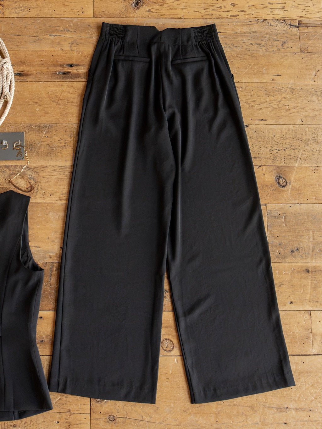 The Cuero Black Vest Set from Savannah Sevens western life{&}style is displayed laid flat on a wooden floor, featuring black high waisted wide leg pants with an elastic waistband paired with a matching black sleeveless top. Crafted from matte satin fabric, the pants provide a relaxed fit and include two back pockets, creating a sophisticated coordinated set.