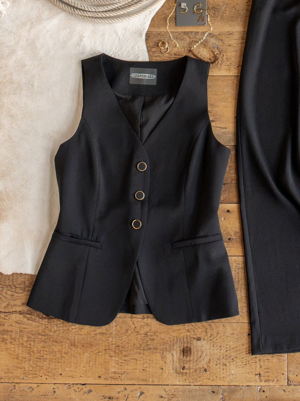 The Cuero Black Vest Set from Savannah Sevens western life{&}style, crafted from matte satin fabric, is displayed on a rustic wooden surface. The tailored vest, featuring three gold buttons, is paired with high waisted wide leg pants and styled alongside a white rug, rope, gold jewelry, and a small ruler.