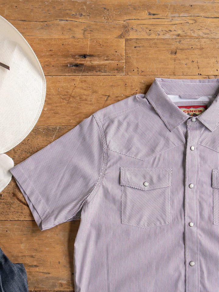 The Cutler Cinch Men's Pinstripe Pearl Snap by Savannah Sevens western life{&}style is laid flat on a wooden surface beside the brim of a white hat.
