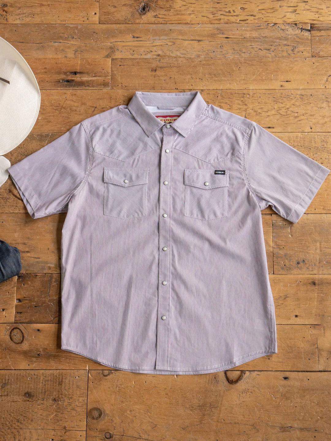 The Cutler Cinch Men's Pinstripe Pearl Snap by Savannah Sevens western life{&}style, a short-sleeve light purple shirt with pearl snaps and two chest pockets, is laid flat on wood beside a white cowboy hat and denim jeans.