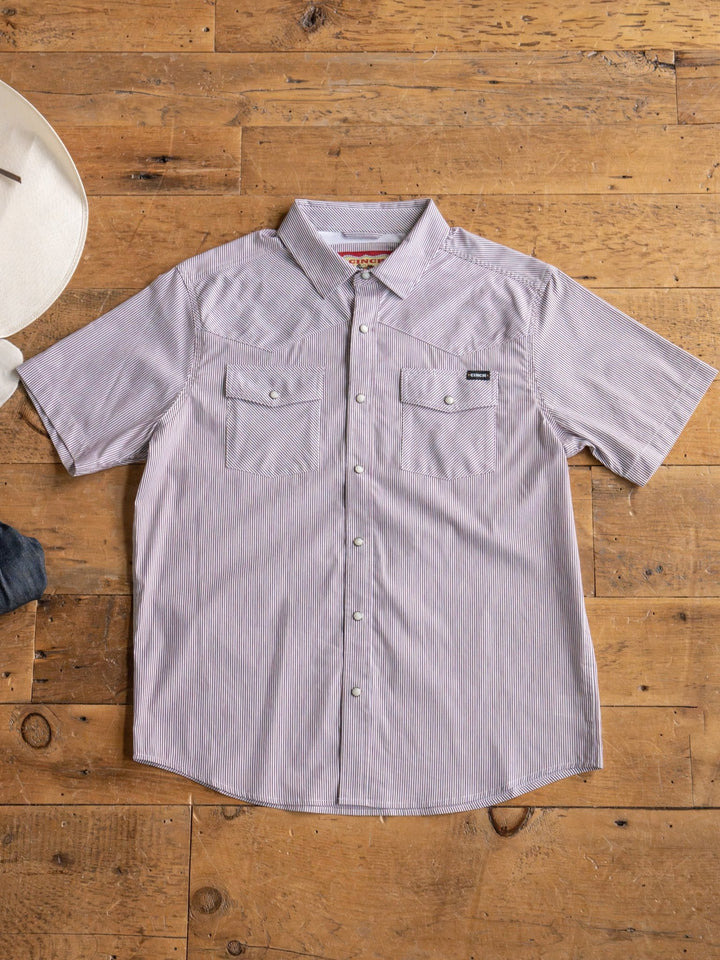 The Cutler Cinch Men's Pinstripe Pearl Snap by Savannah Sevens western life{&}style, a short-sleeve light purple shirt with pearl snaps and two chest pockets, is laid flat on wood beside a white cowboy hat and denim jeans.