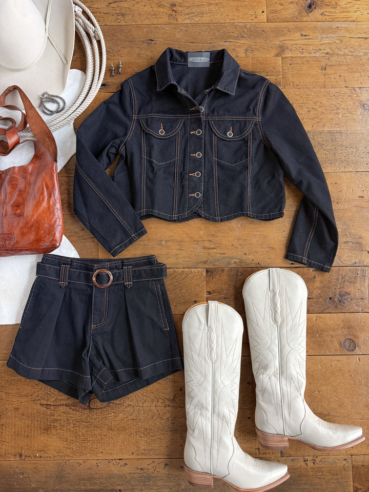 Showcase your Western-inspired look with the Dacoma Black Denim Set from Savannah Sevens western life{&}style, featuring a black denim jacket and matching high-waisted shorts. Complete the outfit with white cowboy boots, a brown leather shoulder bag, a coiled rope, and a white cowboy hat, all arranged on a rustic wooden surface.