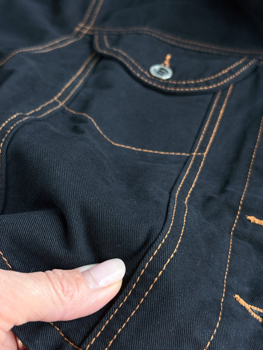 A close-up of a person’s hand pinching the fabric of the Dacoma Black Denim Set by Savannah Sevens western life{&}style, showcasing the rich orange stitching, front pocket, and signature metal button—an essential piece from this stylish women’s denim collection.