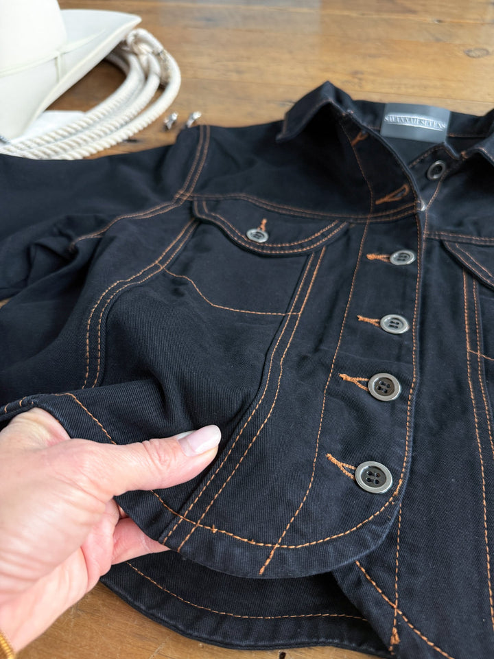 A hand holds the hem of the Dacoma Black Denim Set by Savannah Sevens western life{&}style, featuring orange stitching and gray buttons on black denim, displayed on a wooden surface near a coiled rope and a white object. This stylish piece is part of a women’s denim set.