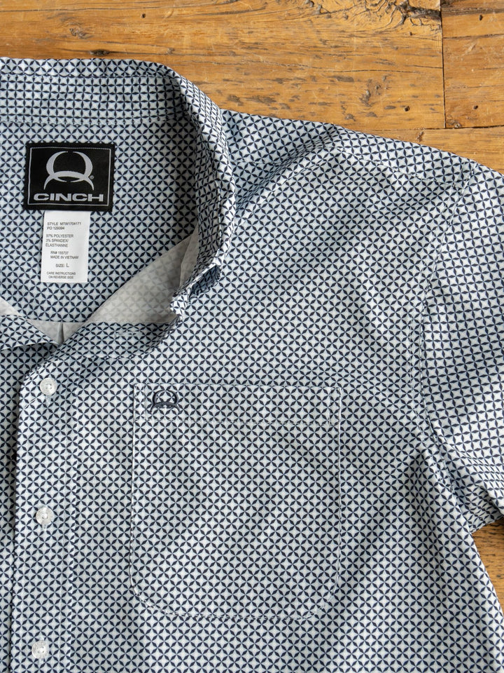 The Dade Cinch Men's Navy Southwest Button Up by Savannah Sevens western life{&}style features a geometric blue and white pattern, front pocket, collar with visible label, UV defense, and is displayed on a wooden surface.