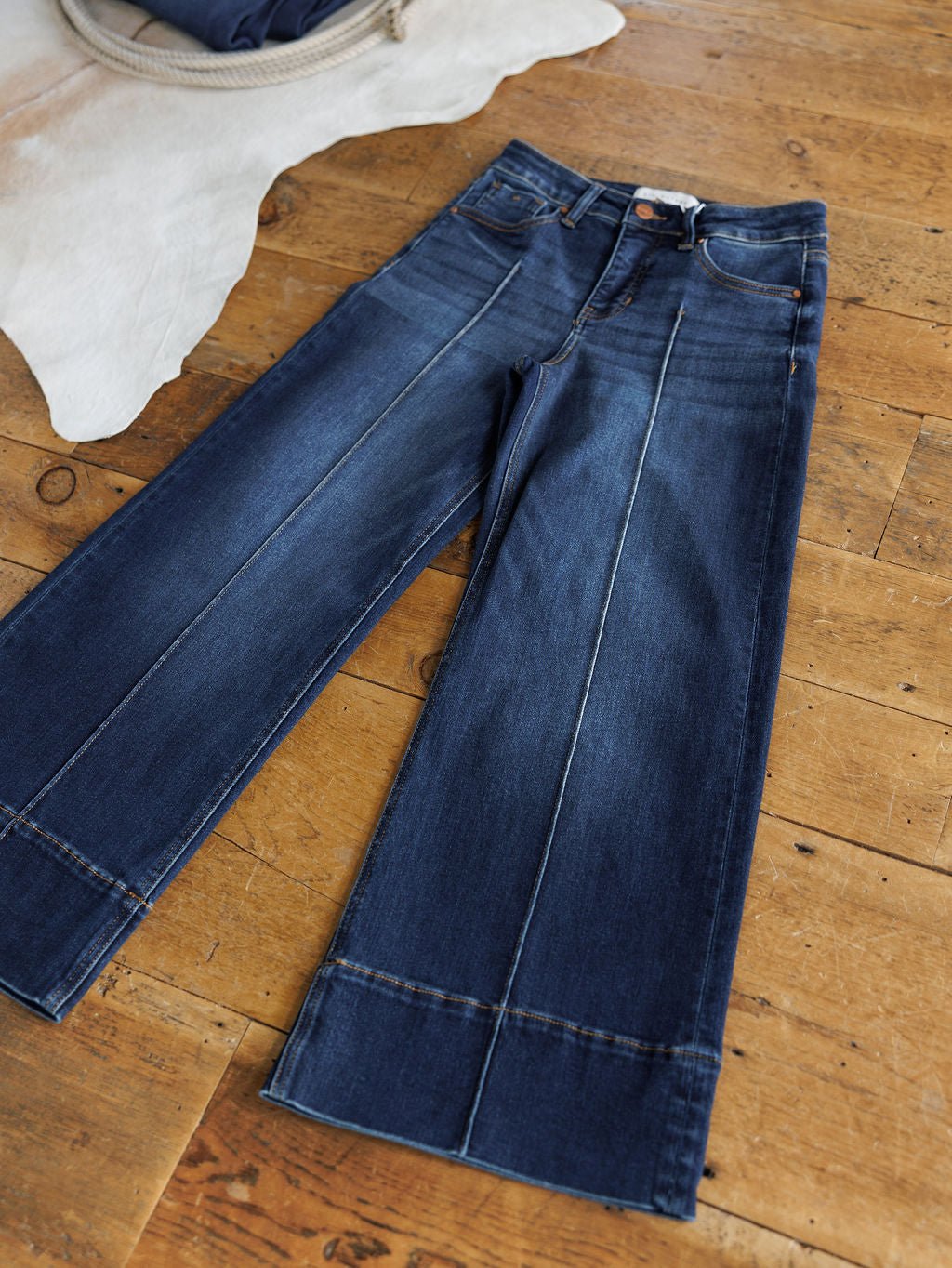 The Dakota High Rise Wide Leg Crop Jean by Savannah Sevens western life{&}style is laid flat on a wooden floor near a white fur rug, showcasing prominent seam stitching, tummy control, button closure, and front and back crease details.