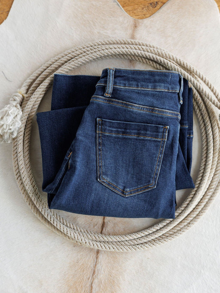 A pair of Savannah Sevens western life{&}style’s The Dakota High Rise Wide Leg Crop Jean, featuring tummy control and a dark wash, is folded inside a coiled rope on a light, textured animal-hide-like surface.