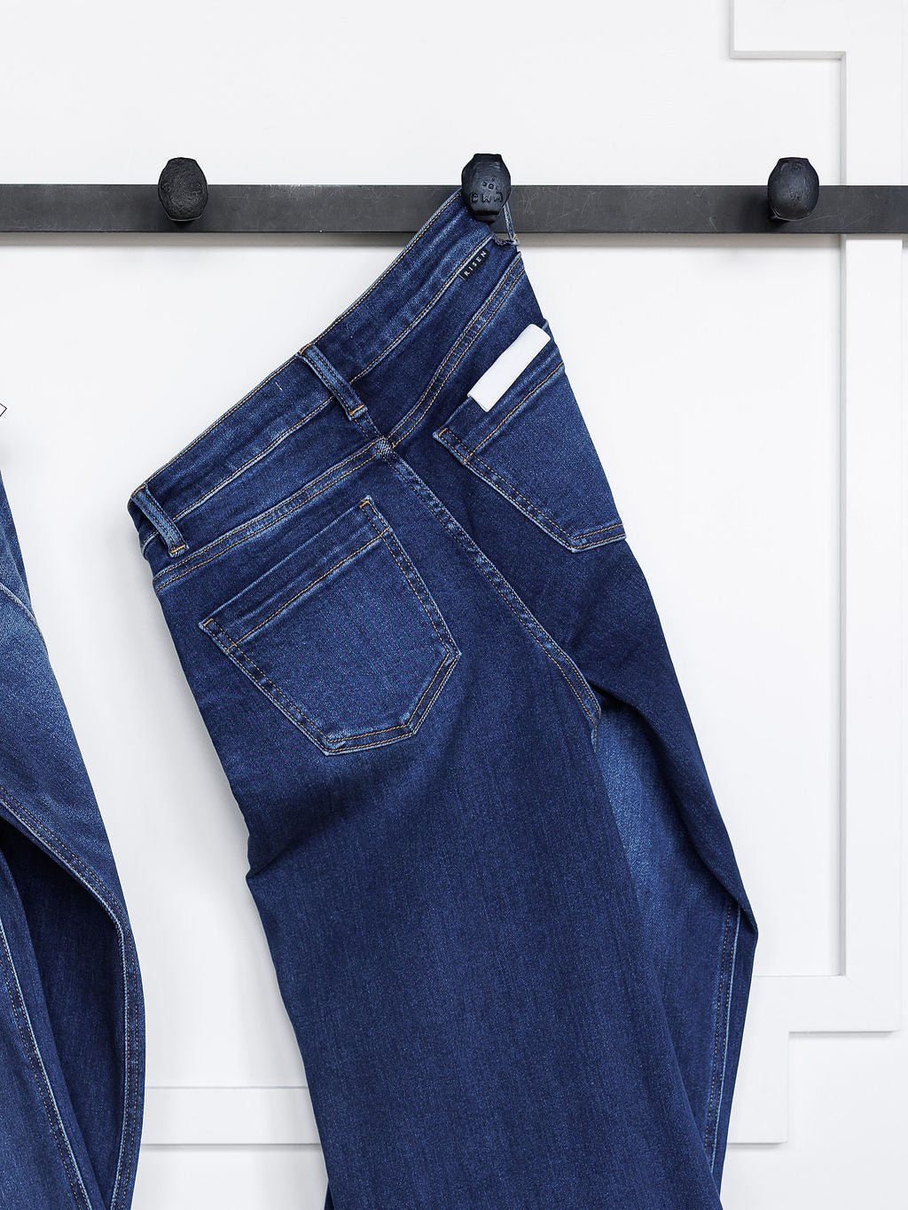 The Dakota High Rise Wide Leg Crop Jean by Savannah Sevens western life{&}style in a dark wash hangs from a black wall hook, with a white item peeking from the back pocket against a white wall.