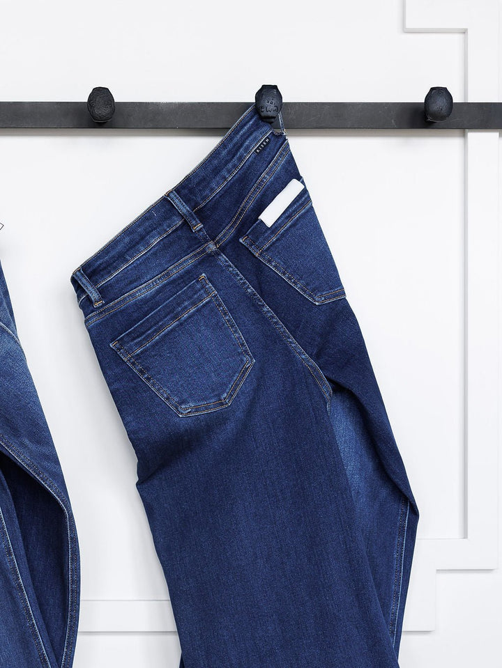 The Dakota High Rise Wide Leg Crop Jean by Savannah Sevens western life{&}style in a dark wash hangs from a black wall hook, with a white item peeking from the back pocket against a white wall.