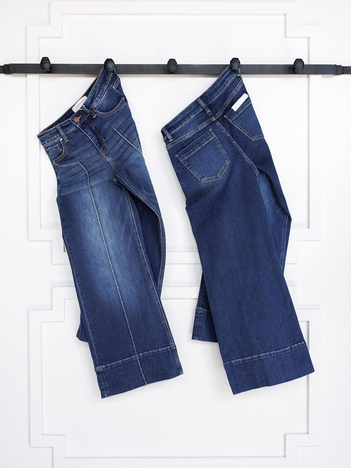 Two pairs of The Dakota High Rise Wide Leg Crop Jeans by Savannah Sevens western life{&}style, featuring dark wash, visible stitching, wide leg crop fit, and stylish tummy control, hang side by side on a black rod against a textured white wall.