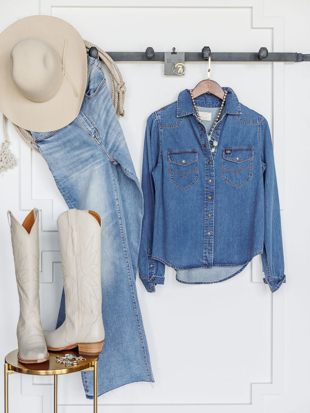 The Dalles by Savannah Sevens western life{&}style—a beige cowboy hat, women's relaxed fit denim jeans, and a western yoke denim shirt—hang on a black rack as white cowboy boots rest on a gold table against a clean white wall.