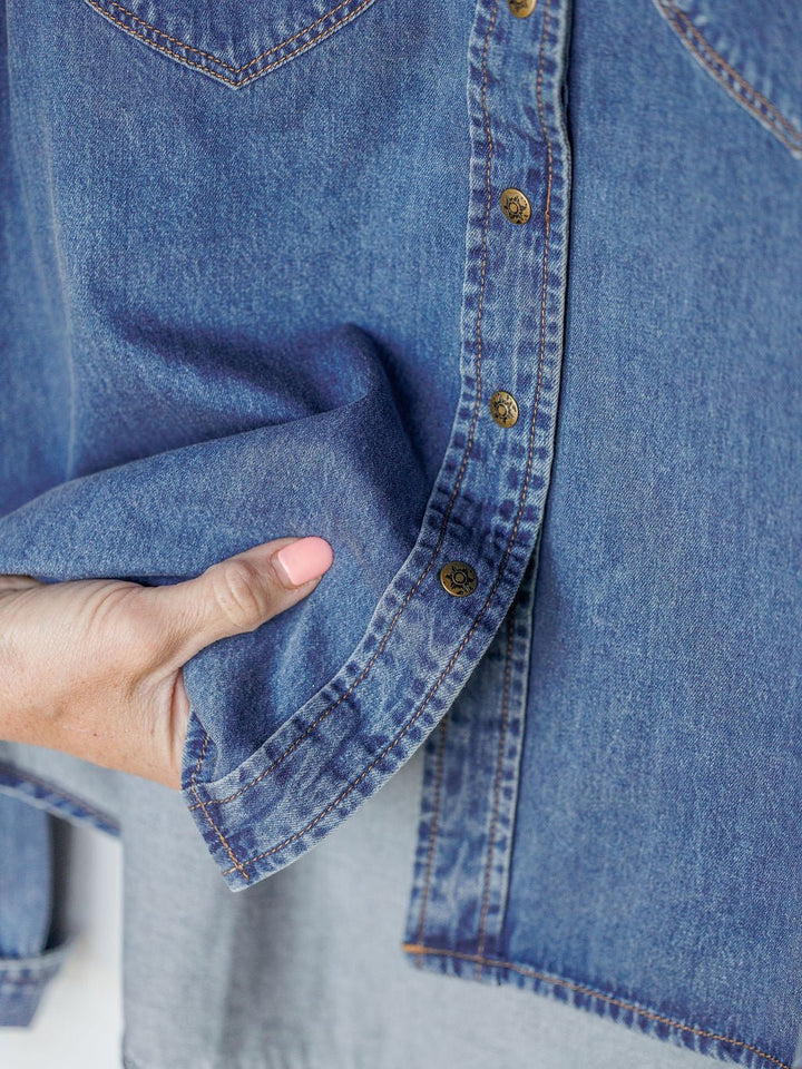 A person places their hand at the hem of The Dalles women’s relaxed fit denim shirt by Savannah Sevens western life{&}style, featuring brass buttons and visible stitching.