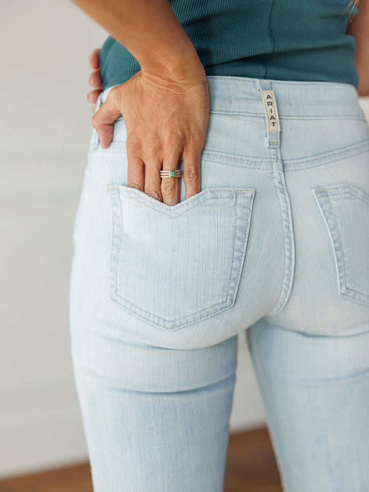 Close-up of a person wearing The Dani Perfect Rise Trouser Jean *Exclusive* by Savannah Sevens western life{&}style, with one hand on their back pocket, teal top, and silver ring. Label on waistband reads AGAIN.