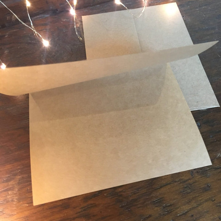 The Darlin' Christmas Card by Savannah Sevens western life{&}style is shown open with its matching envelope on a wooden surface, ready for your personal holiday message. Soft string lights add a festive touch in the background.