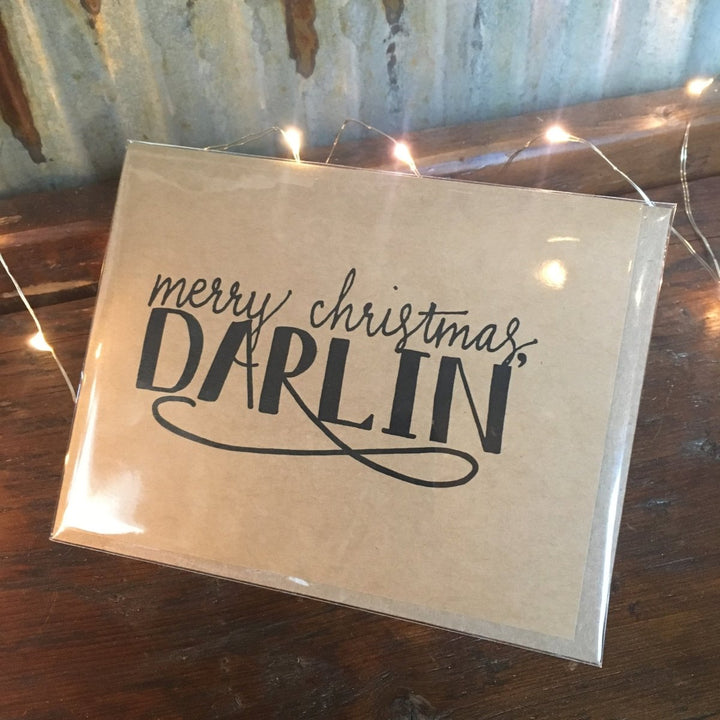 The Darlin' Christmas Card by Savannah Sevens western life{&}style features black script reading "merry christmas, DARLIN" and is displayed on a wooden surface with string lights against a corrugated metal wall.